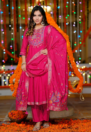 A woman modeling a bright pink traditional Indian outfit with matching dupatta, decorated with floral embroidery and mirror work, set against a festive background with marigold garlands and multicolored string lights.