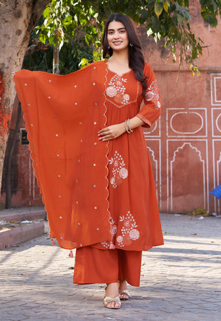 Woman in an orange traditional outfit with white embroidery standing in front of a pink architectural structure.