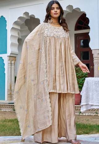 A woman models a traditional beige three-piece salwar kameez suit with a matching embroidered dupatta, set against an architectural background with arches.