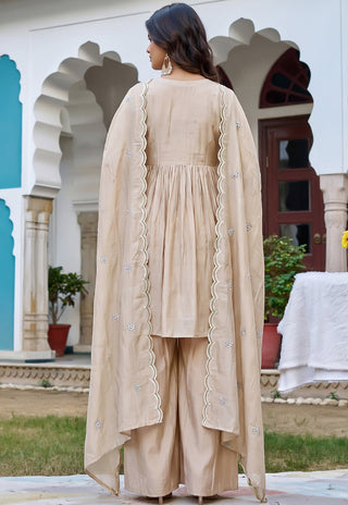 A woman viewed from behind wearing a beige traditional outfit with a long draped jacket featuring scalloped edges and embroidered details, set against an outdoor architectural background with arches.