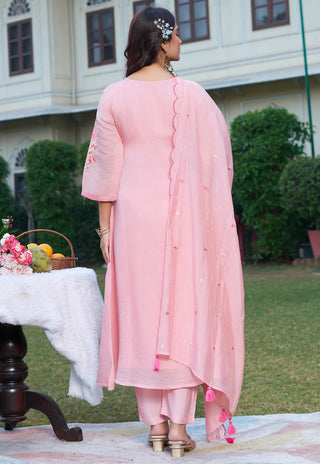 Woman in a pink traditional outfit standing outdoors with a table and flowers in the background.