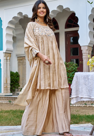 A woman models a beige traditional South Asian outfit with a heavily embroidered yoke and wide sleeves, paired with matching pleated palazzo pants, set against an outdoor architectural background with arches.