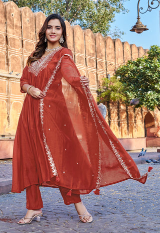 A woman models a rust-red Indian ethnic suit set with a matching dupatta, featuring silver embroidery, standing outdoors in front of a sandstone wall.