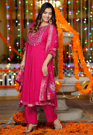 A woman models a bright magenta pink ethnic salwar kameez suit with silver embroidery, matching pants, and a dupatta, posed in front of a festive background decorated with marigold garlands and colorful string lights.