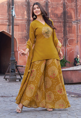 A woman models a mustard yellow ethnic three-piece salwar kameez set with intricate floral embroidery and matching wide-leg trousers in an outdoor setting with a historic terracotta-colored wall.