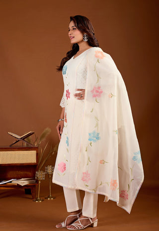 A woman models a traditional white salwar kameez suit with floral embroidery. The set includes a long-sleeved kurta with embroidered details, matching white trousers, and a sheer white dupatta decorated with pink, blue, and peach floral motifs.