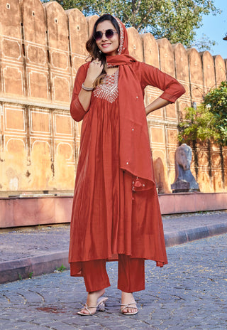 A woman models a rust-orange Indian ethnic wear set consisting of a kurta, matching trousers, and a dupatta, featuring a silver embroidered neckline, set against a backdrop of a stone wall and cobblestone street.