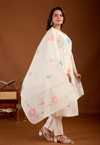 A woman models a traditional white three-piece salwar kameez set with delicate pink and blue floral embroidery, set against a warm brown studio background.