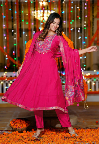 A woman modeling a bright pink traditional Indian salwar kameez suit with matching dupatta, decorated with intricate embroidery on the chest, standing on a floor covered with orange marigold petals in front of a festive backdrop with hanging lights and garlands.