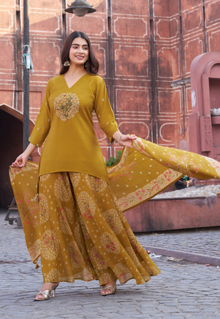 A woman modeling a mustard yellow ethnic salwar kameez set with a matching printed dupatta, standing outdoors in front of a reddish-brown stone wall.