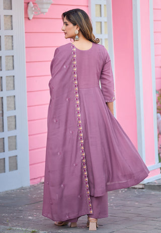 A woman wearing a dusty purple three-piece traditional outfit with embroidered details, standing in front of a pink building.