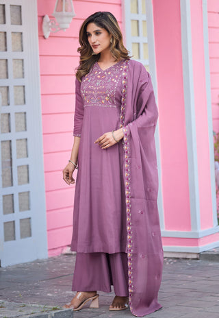 A woman models a three-piece lavender purple ethnic suit consisting of a long kurta with floral embroidery, matching wide-leg trousers, and a dupatta, set against a pink building background.