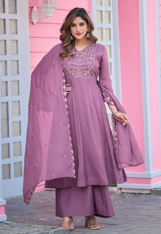 A woman models a three-piece dusty purple anarkali suit with matching palazzo pants and a sheer dupatta, featuring gold and pink floral embroidery on the bodice and sleeve borders, set against a pink building background.