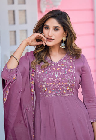 A woman wearing a lavender purple embroidered Anarkali suit with a matching dupatta, featuring colorful floral embroidery on the bodice and sleeves, set against a pink and white background.