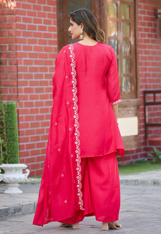 A woman wearing a bright red traditional outfit with matching pants and a draped scarf featuring white floral embroidery, standing outdoors in front of a red brick building.