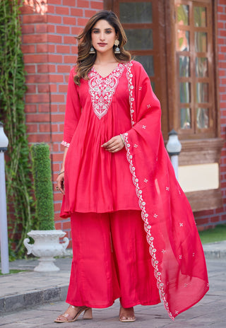 A woman modeling a bright pinkish-red three-piece traditional outfit with matching wide-leg pants and a sheer embroidered dupatta, standing outdoors in front of a brick building.
