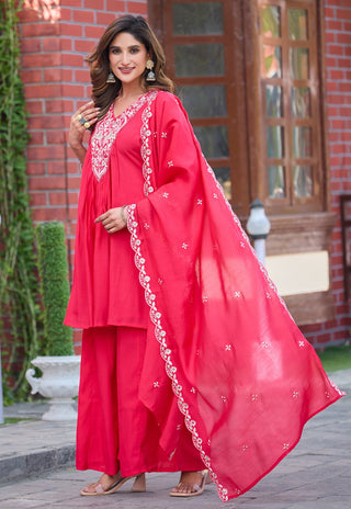A woman modeling a bright pinkish-red traditional outfit with a matching sheer dupatta, featuring white embroidered details on the neckline and along the dupatta border, set against a brick building background.