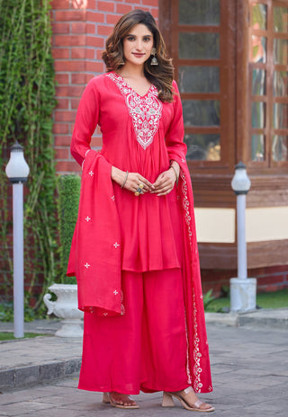 A woman models a bright pink-red traditional Indian outfit with a matching dupatta, featuring white floral embroidery on the neckline and along the dupatta borders, set against an outdoor background of a brick building and greenery.