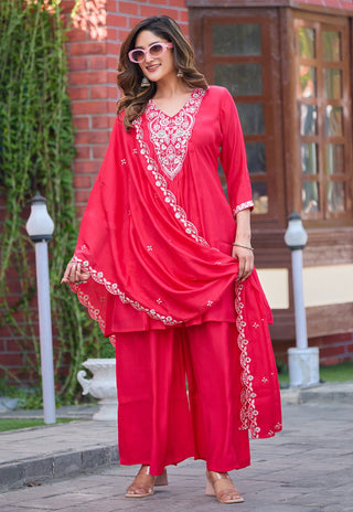 A woman modeling a bright red Indian ethnic wear set with white embroidery, featuring a kurta, wide-leg pants, and a dupatta, standing outdoors in front of a brick building.