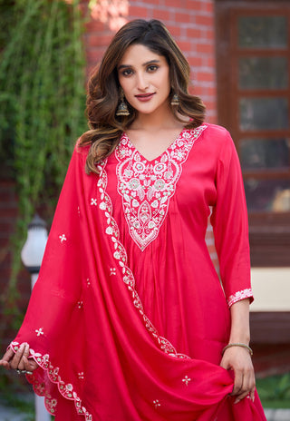 A woman wearing a bright pink-red traditional embroidered kurta set with a matching dupatta, standing outdoors in front of a brick wall.