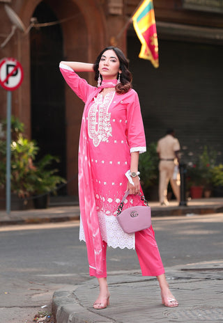 A woman stands on a city street wearing a bright pink traditional outfit with white embroidery and white lace trim, holding a pink handbag.