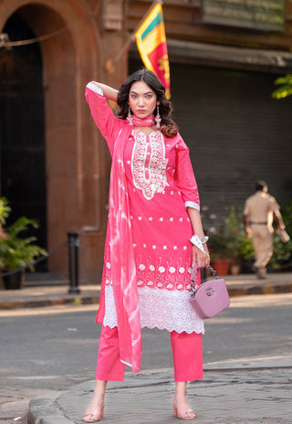 A woman modeling a bright pink traditional Indian salwar kameez set with white lace embroidery, holding a matching pink handbag in an outdoor street setting.