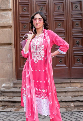 A woman models a bright pink traditional outfit with white floral embroidery, featuring a matching sheer pink dupatta, posed in front of a large brown wooden door.