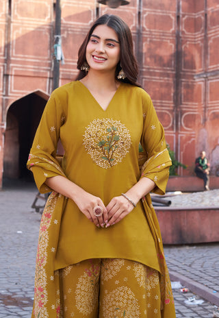 A woman wearing a mustard yellow ethnic three-piece salwar kameez suit with floral embroidery, standing in front of a historic red sandstone building.