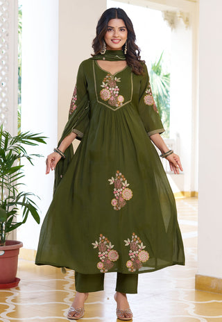 Woman wearing a green embroidered outfit in a bright room with plants.