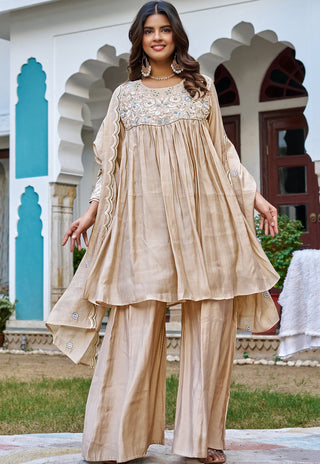 A woman models a beige three-piece traditional outfit with a heavily embroidered yoke, wide sleeves, and matching wide-leg trousers, set against an outdoor architectural background with arches.