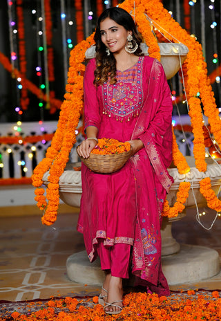 A woman wearing a bright pink traditional outfit with embroidered neckline details and a matching dupatta, standing indoors in front of a stone structure decorated with orange marigold garlands and multicolored string lights. She holds a basket filled with orange marigold flowers.