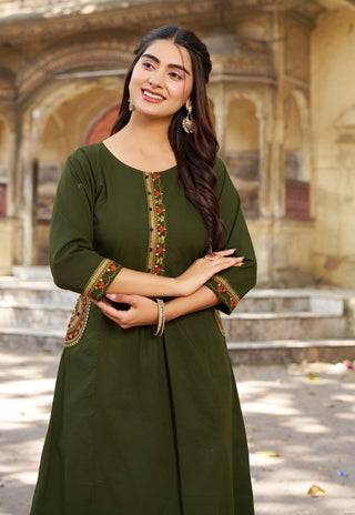 Woman in a green traditional outfit with intricate designs standing in front of an architectural background.