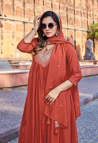A woman wearing a rust-orange traditional Indian salwar kameez suit with a matching dupatta, silver embellished chest embroidery, and sunglasses, posing outdoors in front of a historic stone wall.