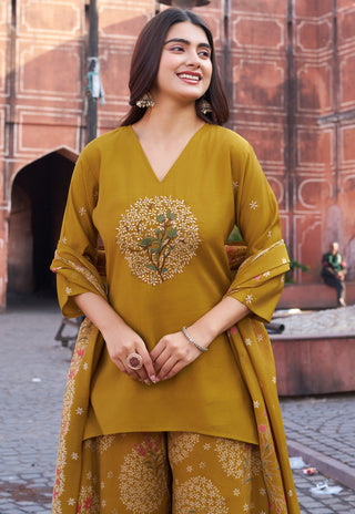 A woman wearing a mustard yellow ethnic three-piece suit with floral embroidery, standing in front of a historic red sandstone wall.