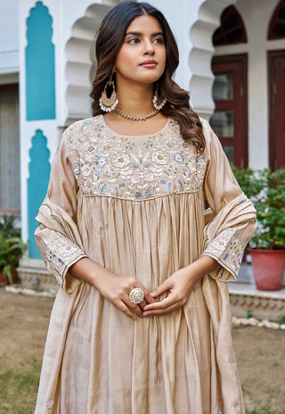 A woman wearing a beige embroidered traditional dress with a heavily decorated yoke and sleeve cuffs, standing outdoors in front of a white building with arches and teal accents.