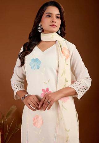 A woman models a cream-colored three-piece salwar kameez suit set with floral embroidery. The set includes a kurta with blue and pink floral motifs, matching embroidered dupatta, and a white embroidered border.