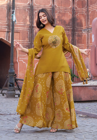 A woman modeling a mustard yellow ethnic three-piece suit with wide-leg patterned pants and a matching dupatta, standing outdoors in front of a reddish-brown stone wall.