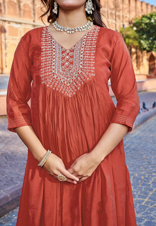 A woman wearing a rust-orange cotton kurta with intricate silver mirror work embroidery around the V-neckline, styled with traditional Indian jewelry in an outdoor setting.