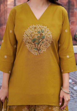 A woman wearing a mustard yellow V-neck kurta with a central floral embroidery motif and small embroidered star details on the sleeves, paired with matching patterned trousers.