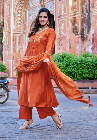 A woman models a rust-orange traditional Indian salwar kameez suit with matching dupatta in front of a historic arched stone gateway. The outfit features an embroidered kameez top, wide-leg pants, and a flowing dupatta.