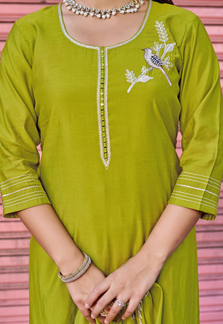 A woman wearing a lime green kurta with white embroidery of a bird and branch on the chest, and white striped detailing on the cuffs. She is wearing silver jewelry, including a beaded necklace and bangles, against a pink corrugated metal background.