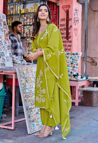 A woman modeling a bright olive green traditional Indian salwar suit set with white embroidery and a matching dupatta, standing in a street market setting.