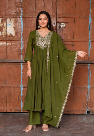 A woman models an olive green traditional Indian outfit consisting of a kurta with silver embroidery, matching palazzo pants, and a dupatta, posed in front of a rustic brown wooden door.