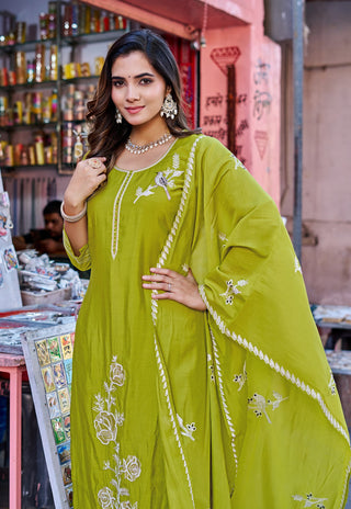 A woman modeling a bright olive green traditional outfit with white floral embroidery and a matching embroidered dupatta, posed in front of a colorful shop background.