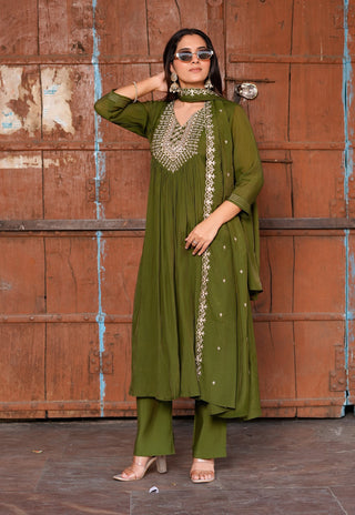 A woman models an olive green ethnic salwar kameez suit with silver embroidery, standing in front of a rustic brown wooden door.