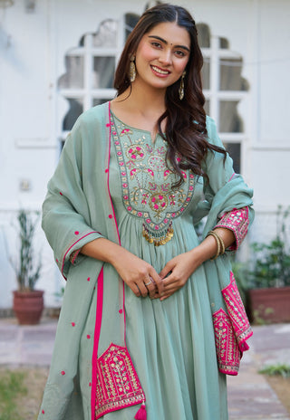 A woman wearing a sage green traditional outfit with vibrant pink and gold embroidery, standing in front of a white building with arched windows.