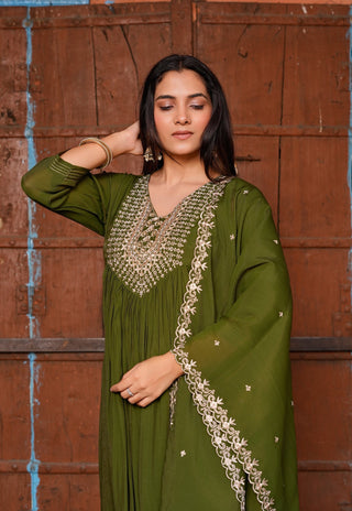 A woman wearing an olive green traditional outfit with intricate gold embroidery, standing in front of a rustic brown wooden door.