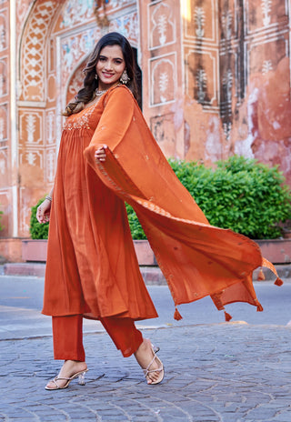 A woman modeling a rust-orange traditional Indian salwar kameez suit with matching pants and a flowing dupatta, set against a historic stone wall with green bushes.
