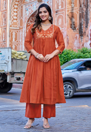 A woman models a rust-orange kurta set with matching wide-leg pants, standing outdoors in front of a historic stone wall and street scene.