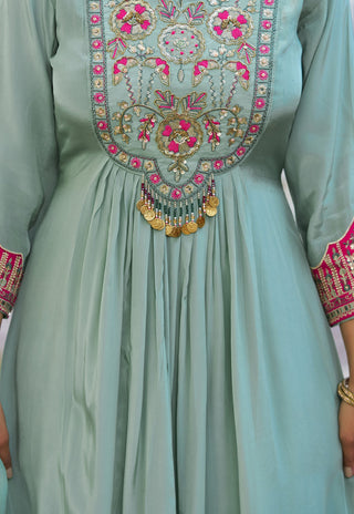 A close-up of a sage green traditional dress with intricate gold, pink, and red embroidery on the chest and sleeve cuffs, featuring a gold coin tassel.